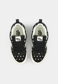 Musta mokkanahkakengät, joissa on kukkakoristeiset niitit, valkoiset pohjat ja mustat nauhat. Vans-logo näkyvissä kengän kielessä. Litteä muoto, pyöreä varvastyppi.