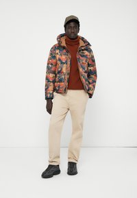 Doudoune en camouflage multicolore avec un col montant, associée à un pull col roulé rouille, un pantalon beige et des baskets noires, se tenant devant un fond blanc.