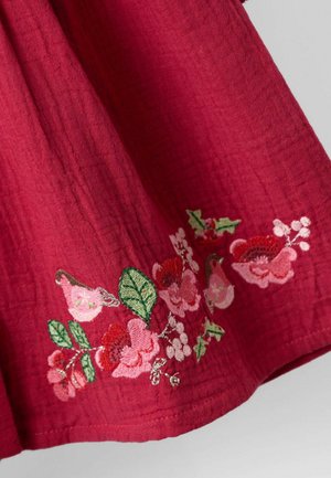 Roter Stoff mit rosa und grüner floralem Stickerei entlang der Kante, die detaillierte Blütenblätter und Blätter zeigt.