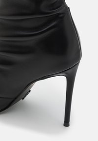 Schwarzer High-Heel-Schuh mit glatter, glänzender Lederoberfläche, ausgestattet mit einem zulaufenden Stiletto-Absatz und dezenten, gerafften Stoffdetails.