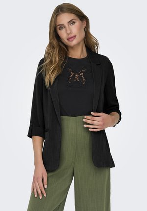 Blazer nero con maniche arrotolate sopra un top nero con un design in pizzo a farfalla, abbinato a pantaloni larghi verdi con texture.