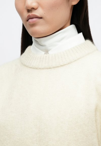 Απαλή μπεζ ζακέτα με ριγωτό λαιμό, επικαλυμμένη πάνω από ένα λευκό πουκάμισο με γιακά. Το ύφασμα φαίνεται μαλακό με λεία υφή.