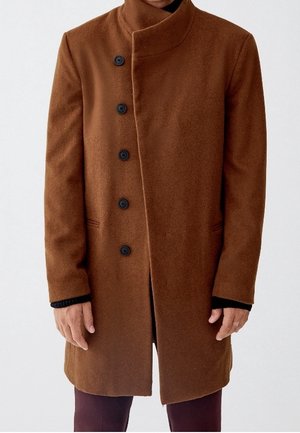 Brązowy wełniany płaszcz z asymetrycznym zapięciem na guziki, stojącym kołnierzem i kieszeniami z rozcięciami, noszony na czarnym, długorękawowym elemencie garderoby.