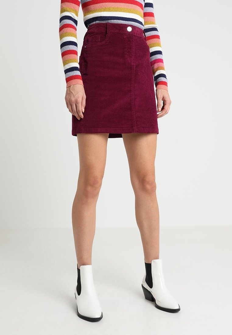 Minifalda de pana burdeos con cierre de botones frontales y dos bolsillos, combinada con botines negros y blancos y una blusa de rayas multicolor.