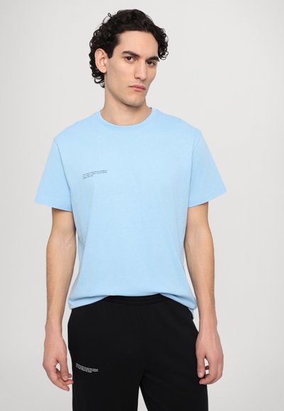 Lys blå bomulds T-shirt med korte ærmer, med en lille trykt tekstdetalje på venstre side. Sammen med sorte bukser.
