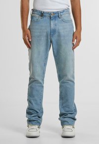 Šviesiai mėlynos džinsinės kelnės su tiesiais kelnių siluetais, turinčios penkis kišenes, diržo kilpas ir mygtuką, derinamos su baltomis sportbačiais.