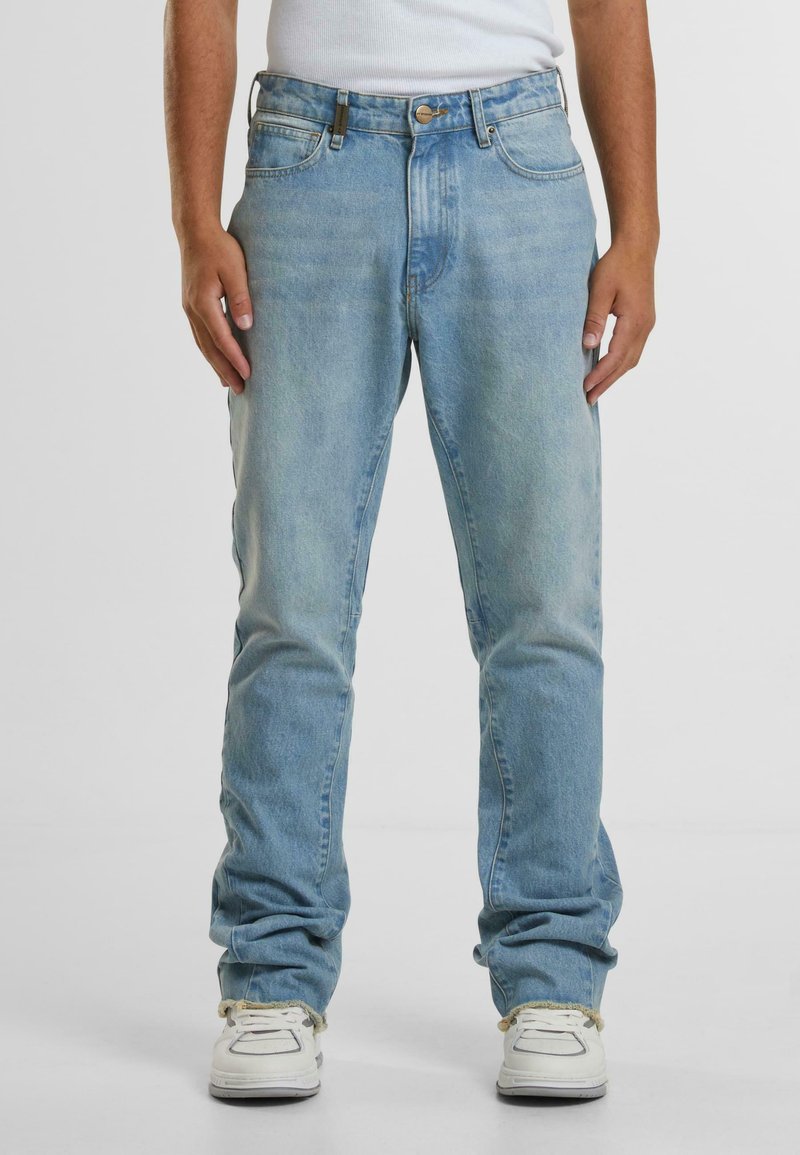 Šviesiai mėlynos džinsinės kelnės su tiesiais kelnių siluetais, turinčios penkis kišenes, diržo kilpas ir mygtuką, derinamos su baltomis sportbačiais.