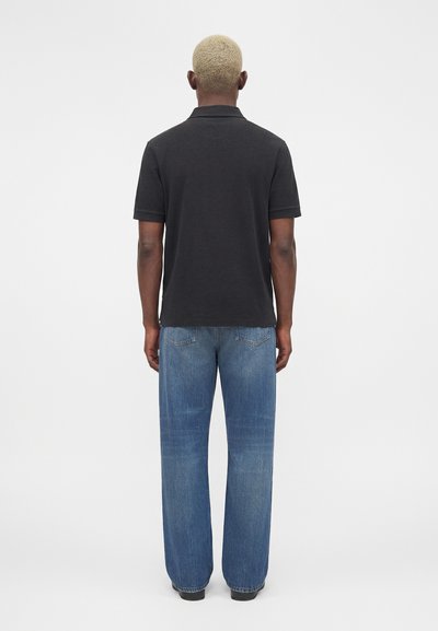 Άτομο με κοντά ξανθά μαλλιά που φοράει μαύρο κοντομάνικο πόλο και φαρδιά μπλε τζιν παντελόνια, στέκεται γυρισμένο με την πλάτη σε απλό φόντο.