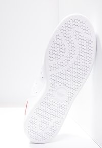 Vit sneakersula med ett bikake-mönster, med en slät yta och en röd accent nära hälen, som visar upp en präglad logotyp.