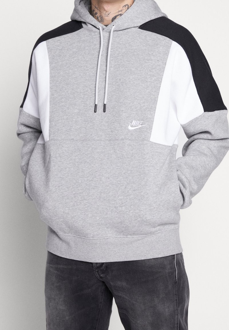 Harmaa huppari, jossa mustia ja valkoisia yksityiskohtia, hupullinen malli, nyörit ja pieni valkoinen logo. Pehmeä rakenne ja rento istuvuus.