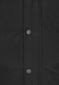 Zwarte jas van gewatteerde stof met een knoopsluiting aan de voorkant voorzien van grote, ronde zwarte knopen. Gladde textuur met zichtbare stiksels en naden.