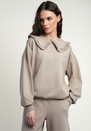 Młoda kobieta ubrana w beżowy strój wypoczynkowy z dużym, falbanianym kołnierzem, patrzy na bok z jedną ręką w kieszeni.