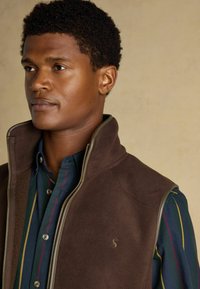 Hnedý fleecový vek s vysokým golierom a zipsom, nosený cez tmavomodrú košeľu s vertikálnymi pruhmi v zelenej, červenej a žltej farbe. Výrazný detail loga.