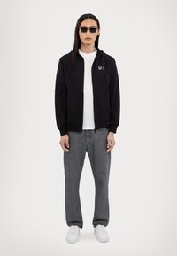 Svart zip-up hoodie, grå t-shirt, grå jeans och vita sneakers. Modell med långt mörkt hår och rektangulära solglasögon. Enkel, avslappnad stil.