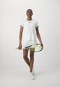 Valkoinen urheilupaita lyhyillä hihoilla ja mustalla logolla, yhdistetty valkoisiin hameisiin. Pelaaja pitää tennismailaa, jossa on keltaisia tennispalloja.