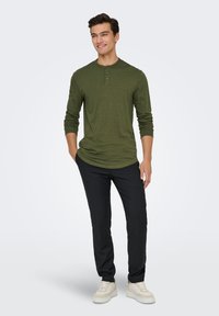 Groen henleyshirt met lange mouwen en een drieknoopssluiting, gecombineerd met zwarte broek en lichtgekleurde sneakers, gedragen door een mannelijke model die staat.