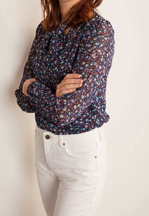 Blusa a stampa floreale in tessuto trasparente e scuro con maniche lunghe e scollo a fiocco, abbinata a jeans bianchi attillati.