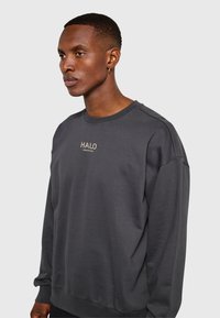 Jonge man met kort haar, gekleed in een donkergrijze trui met de tekst "HALO" op de borst, staand tegen een effen lichte achtergrond.