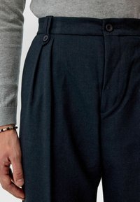 Donkerblauwe op maat gemaakte broek met twee voorplooien, de stofstructuur zichtbaar, voorzien van twee zwarte knopen op de tailleband. Grijze sweaterhengsel zichtbaar.