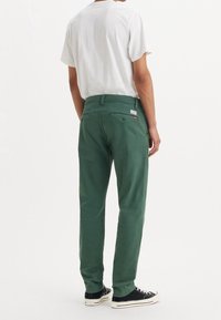 Pantalon en coton vert avec une coupe droite, doté de poches arrière et d'une petite étiquette rouge. Associé à un t-shirt blanc à manches courtes et des chaussures noires.