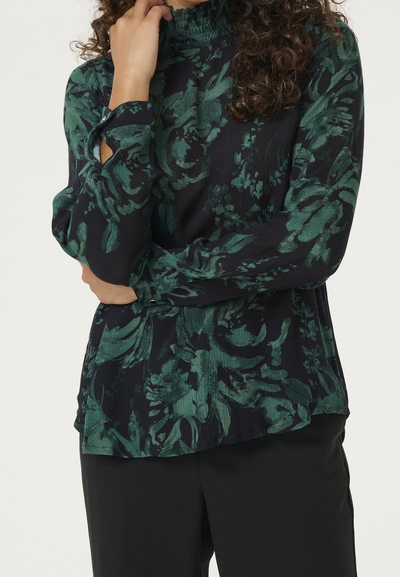 Femme portant un chemisier noir à manches longues avec un motif floral vert et un col montant, associé à un pantalon noir, les bras croisés à la taille.