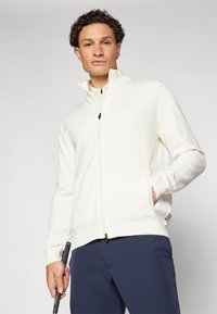 Casaco branco com fecho éclair, feito de um tecido suave, apresentando um colarinho alto, dois bolsos laterais e um logótipo discreto. Usado com calças azul-marinho.