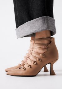 Las botas de tobillo de cuero beige cuentan con múltiples correas horizontales con acentos circulares, puntas afiladas y tacones delgados. Se pueden ver los puños de denim enrollados.