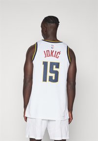 Biely basketbalový dres s modrými a žltými akcentmi, na zadnej strane s menom "JOKIĆ" a číslom "15". Textúrovaný dizajn látky.