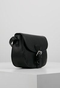 Černá kožená taška přes rameno s klopou a stříbrnou přezkou, vystavená na bílé ploše na šedém pozadí.