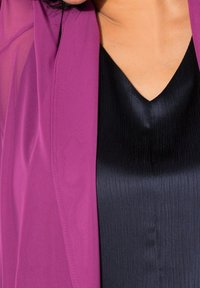 Närbild av en person som bär en lila blazer över en svart V-ringad texturerad topp, vilket visar halslinjen och övre bröstområdet.