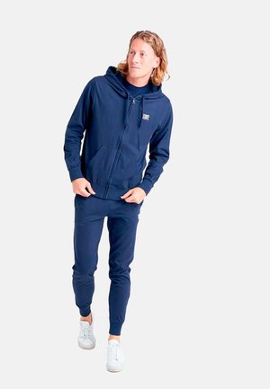 Jeune homme portant un sweat à capuche zippé bleu marine et un jogging assorti avec des baskets blanches, debout et regardant sur le côté.
