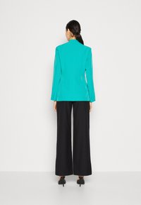 Blazer turquesa con mangas largas, diseño elegante, espalda ajustada, combinado con pantalones de pierna ancha negros y zapatos de tacón, textura suave.