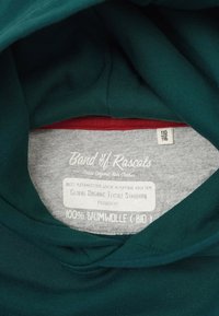 Grön bomullshuvtröja invändigt med en grå etikett som visar vårdingsinstruktioner, certifiering av ekologisk textil och detaljer om tygkomposition.