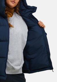 Marineblaue Puffjacke mit Kapuze, ausgestattet mit einer glatten, strukturierten Außenseite und einem grauen Sweatshirt darunter. Reißverschlussdetails sind an der Jacke sichtbar.