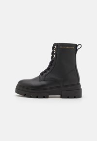 Botte de combat en cuir noir à lacets avec semelle épaisse, tirant au talon et logo doré "Tommy Hilfiger" sur le côté supérieur.