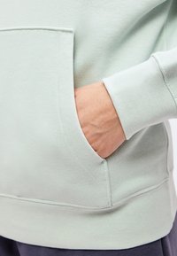 Χέρι εν μέρει μέσα στην μπροστινή τσέπη μιας ανοιχτόχρωμης μπλούζας με ορατές ραφές και ριμπ μανσέτα.