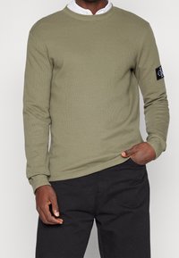 Sweat-shirt à manches longues vert olive avec un tissu texturé, col rond et un patch logo sur la manche gauche. Porté avec un pantalon noir.