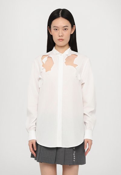 Chemise blanche avec des découpes aux épaules, fermeture à boutons sur le devant et manches longues, accompagnée d'une jupe plissée grise.