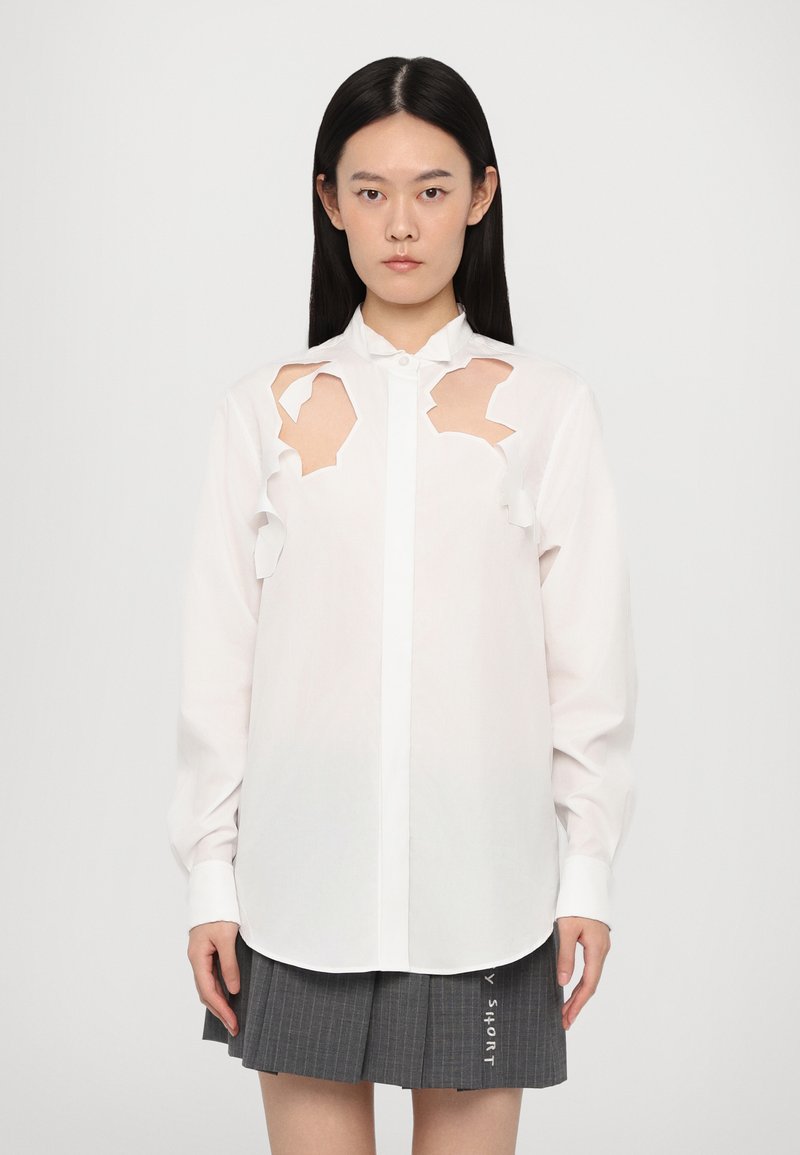 White shirt featuring cut-out details on the shoulders, button-down front, and long sleeves, paired with a gray pleated skirt.