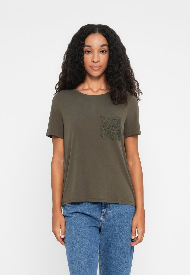 Woman with long curly hair wearing an olive green t-shirt with lace pocket and blue jeans, standing against a white background.