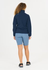 Kvinde med krøllet hår iført en marineblå jakke, lyseblå knælange shorts og sorte sandaler, stående med ryggen til mod en ensfarvet baggrund.