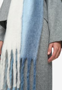 Gradient sjaal met witte, lichtblauwe en donkerblauwe wol, met franjes, gedragen over een gemêleerd grijs kledingstuk.