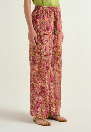 Donna che indossa pantaloni larghi e larghi con motivi floreali astratti rosa, verdi e beige abbinati a sandali gialli e un top verde.