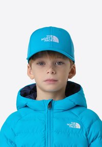 Malé dítě s jasně modrou čepicí The North Face a odpovídajícím modrým péřovým kabátem s kapucí, hledící přímo vpřed na bílém pozadí.