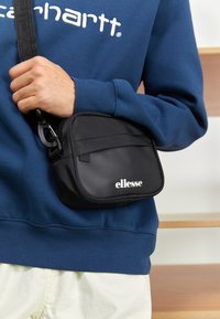 Černá obdélníková crossbody taška s lesklým povrchem, disponující zipovým uzávěrem a bílým logem "ellesse". Nese ji osoba v modré mikině.