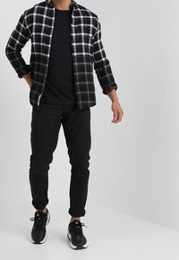 Hombre con camisa de cuadros en blanco y negro sobre camiseta negra, pantalones negros y zapatillas negras, de pie frente a un fondo gris liso.