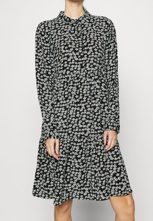 Frau trägt ein schwarzes langärmeliges Kleid mit hellblauem und weißem Blumenmuster, steht mit locker an den Seiten hängenden Händen vor einem schlichten Hintergrund.