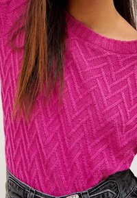 Magenta trui met een visgraatpatroon, voorzien van een wijde halslijn en een zachte textuur. Gepaard met hoge zwarte jeans.