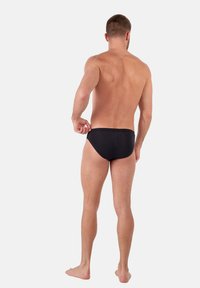 Bañador negro, diseño ajustado, material suave, cintura baja, estilo minimalista, sin patrones visibles ni herrajes, usado por un modelo visto desde atrás.