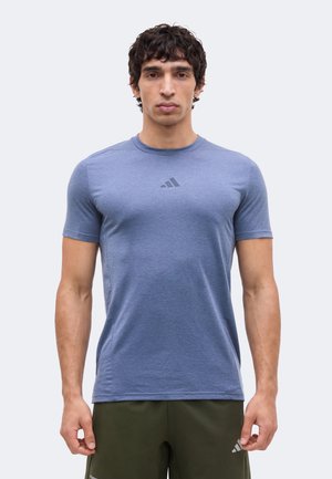 Jeune homme aux cheveux bouclés foncés, portant un t-shirt bleu moulant Adidas et un short vert olive, debout devant un fond clair uni.